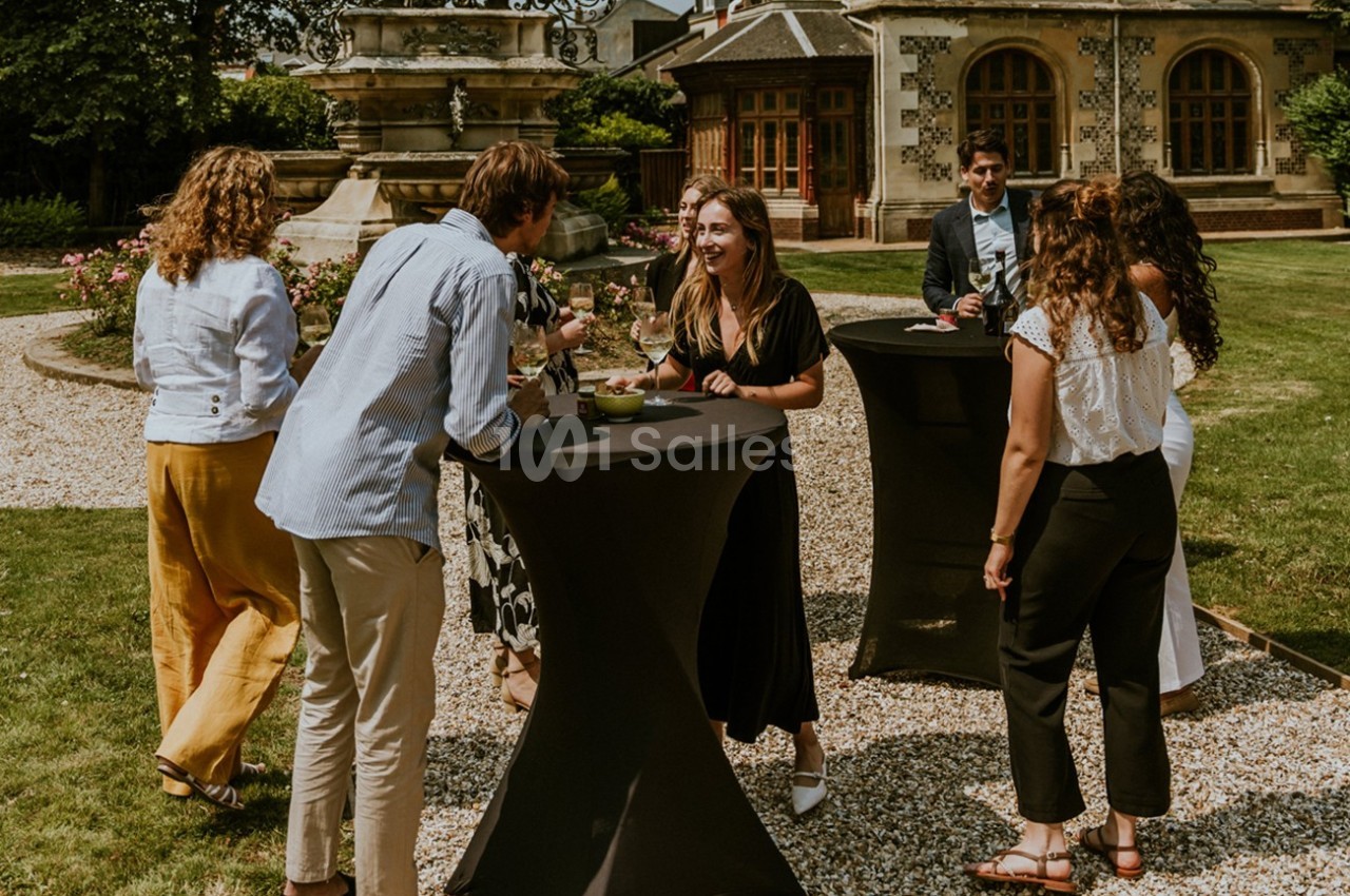 Des personnes discutent autour de tables hautes dans un jardin, près d'une fontaine et d'un bâtiment ancien.