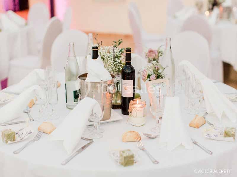Table dressée avec nappes blanches, bouteilles de vin, bougie, fleurs et couverts pour un repas élégant.