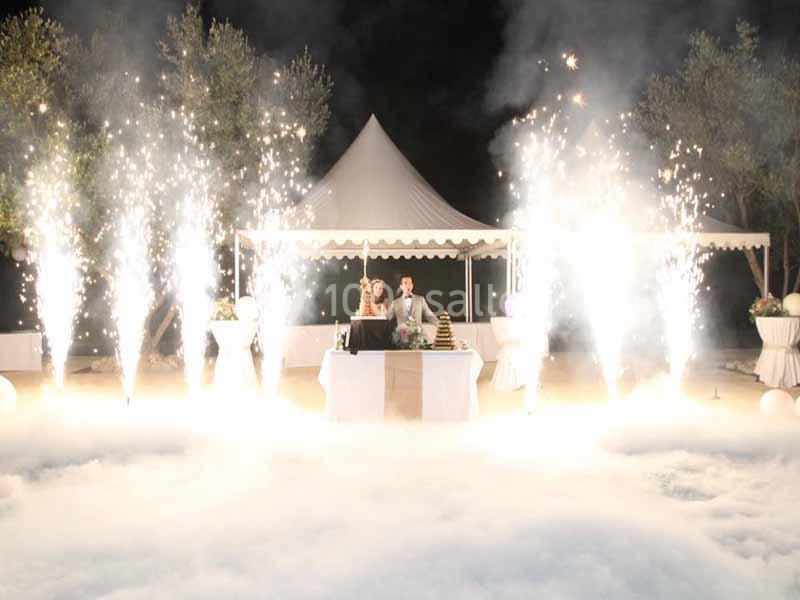 Un couple debout derrière une table décorée, entouré de jets de lumière et de fumée, sous une tente blanche.