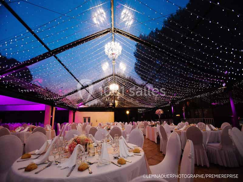 Salle de réception en soirée sous une verrière illuminée, tables rondes dressées avec nappes blanches et décorations…