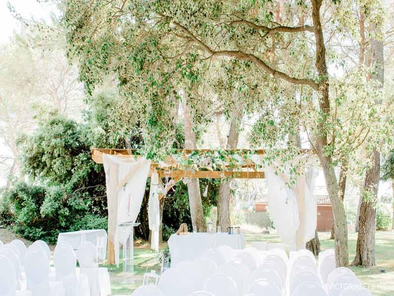 Chaises blanches disposées en rangées devant une arche décorée de voilages et de fleurs dans un jardin ombragé.