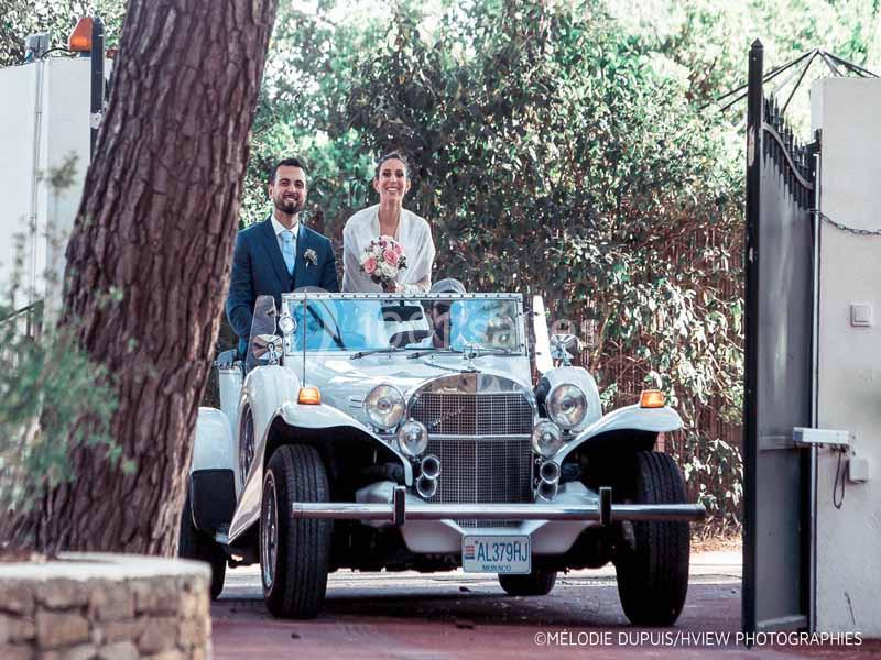 Un couple souriant dans une voiture ancienne décapotable, la mariée tenant un bouquet de fleurs.