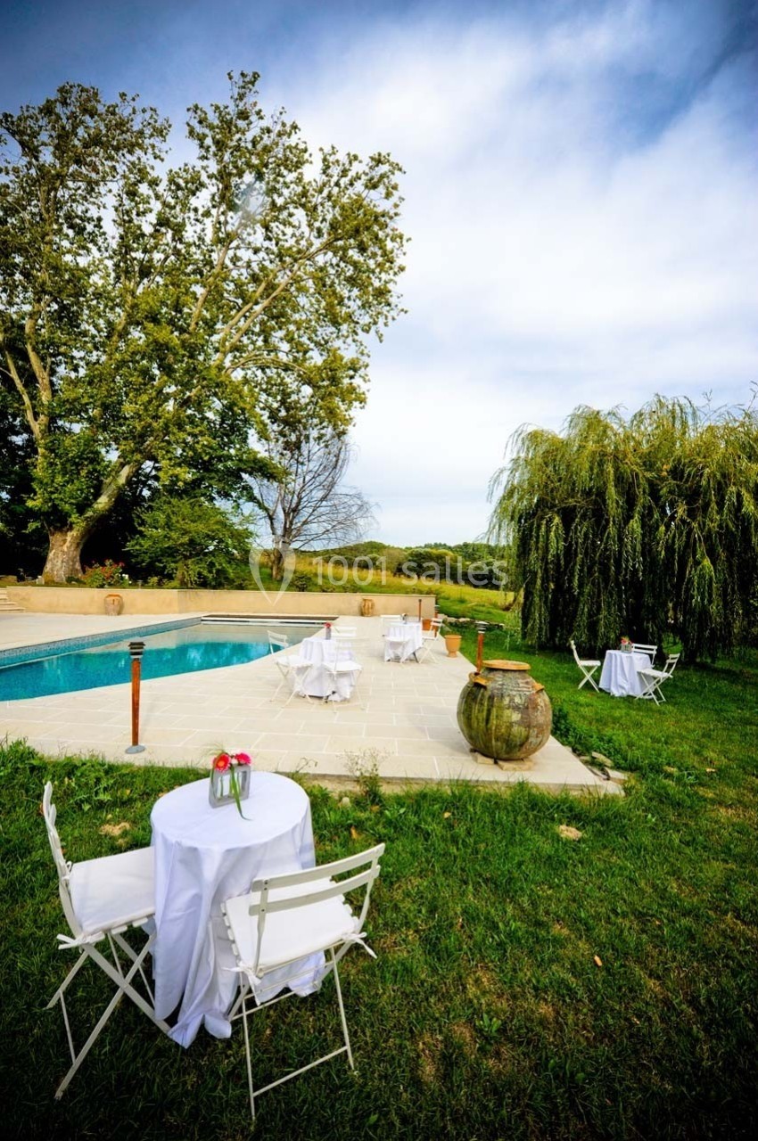 Location salle Ansouis (Vaucluse) - Le Grand Turcan #6 Tables blanches dressées près d'une piscine entourée de verdure et d'arbres sous un ciel légèrement nuageux.