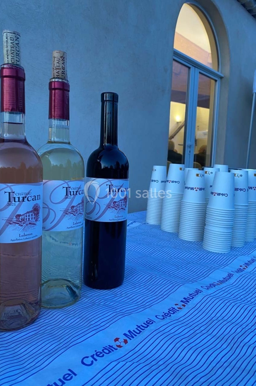 Location salle Ansouis (Vaucluse) - Le Grand Turcan #14 Trois bouteilles de vin et des gobelets en carton disposés sur une table recouverte d'une nappe rayée.