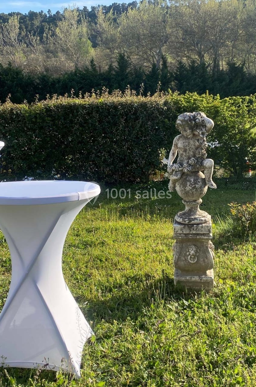 Location salle Ansouis (Vaucluse) - Le Grand Turcan #8 Statue en pierre représentant un enfant assis sur un piédestal, entourée de tables hautes et de verdure.
