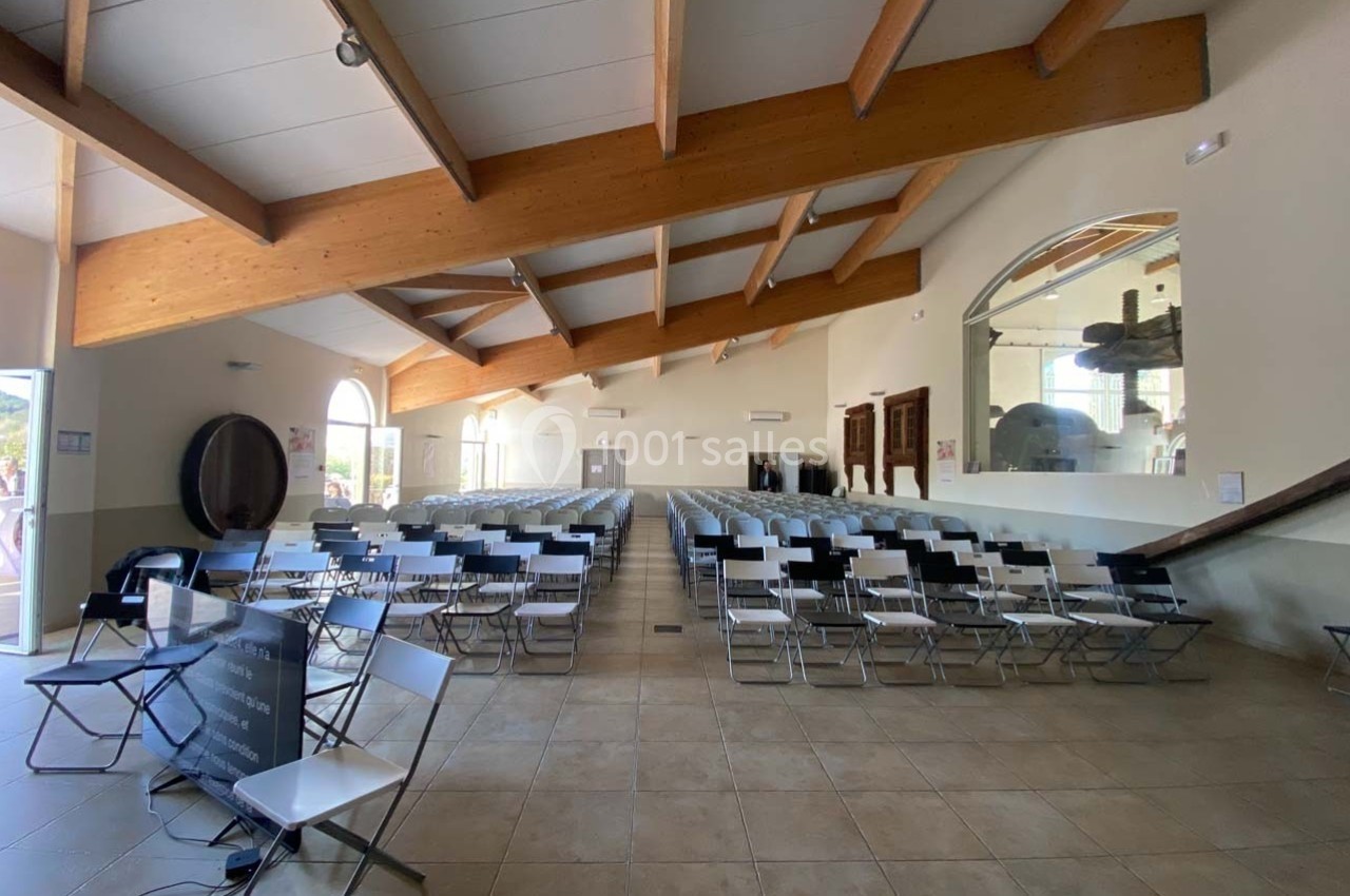 Location salle Ansouis (Vaucluse) - Le Grand Turcan #3 Salle lumineuse avec des rangées de chaises pliantes, plafond en poutres apparentes et grande baie vitrée.
