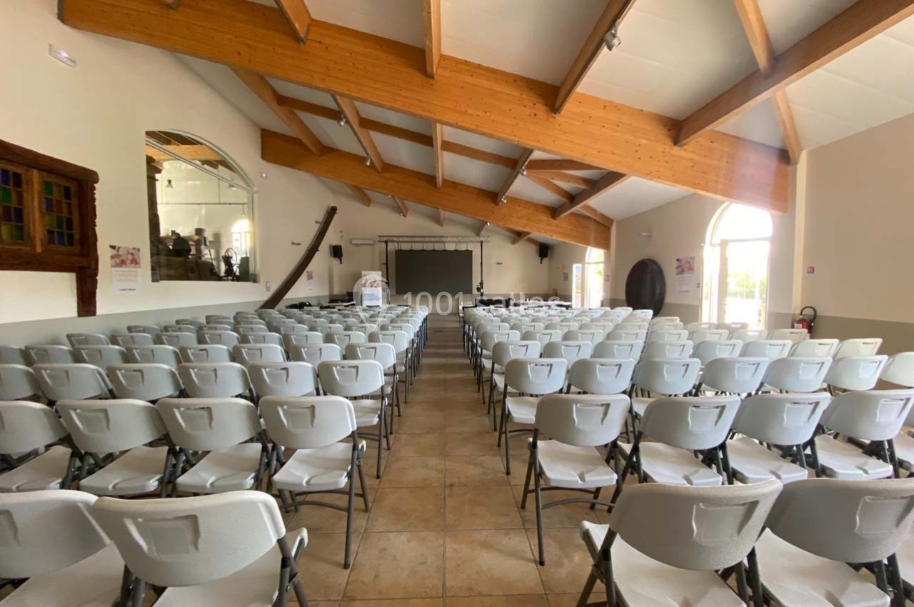 Location salle Ansouis (Vaucluse) - Le Grand Turcan #1 Salle lumineuse avec des rangées de chaises blanches alignées face à un écran et une scène, sous une charpente en bois.