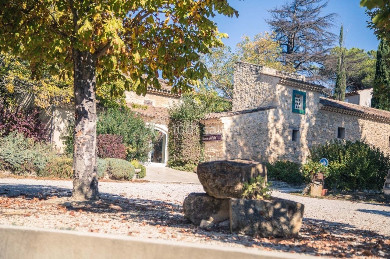 Location salle Ansouis (Vaucluse) - Le Grand Turcan #2 Cour d'une maison en pierre entourée de végétation, avec des arbres aux feuilles automnales et des pierres empilées au…