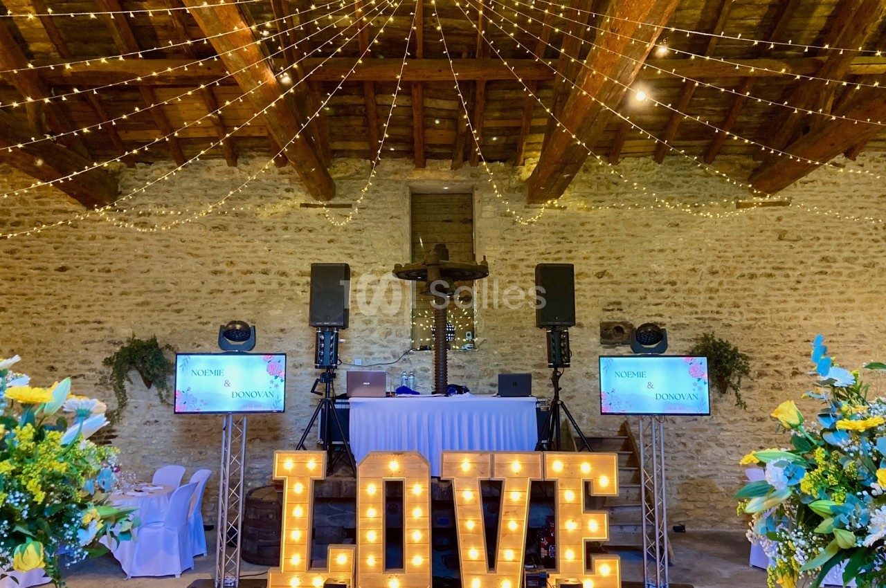 Salle en pierre décorée pour un événement avec guirlandes lumineuses, lettres géantes ’LOVE’ éclairées et équipements audio.