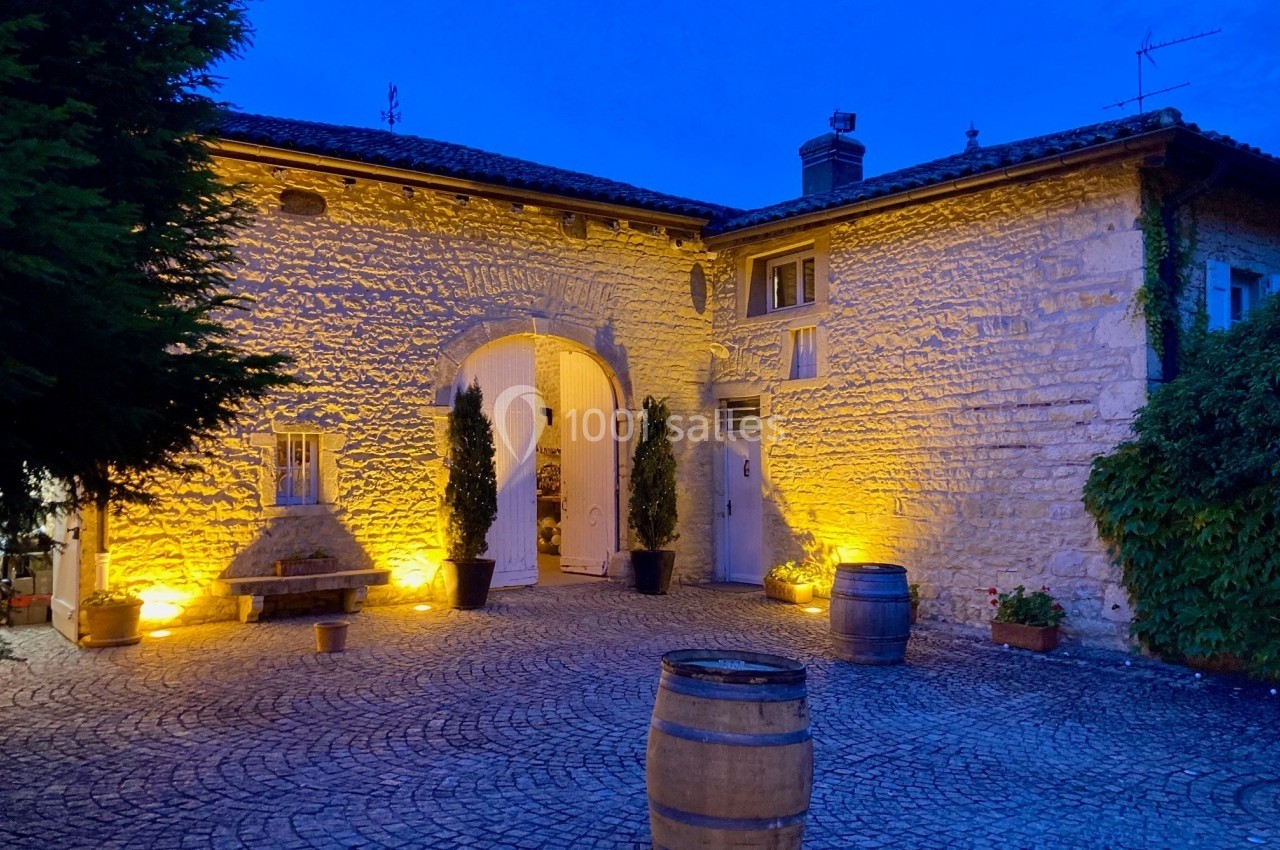 Façade en pierre d'un bâtiment éclairé la nuit, avec des tonneaux et une cour pavée au premier plan.