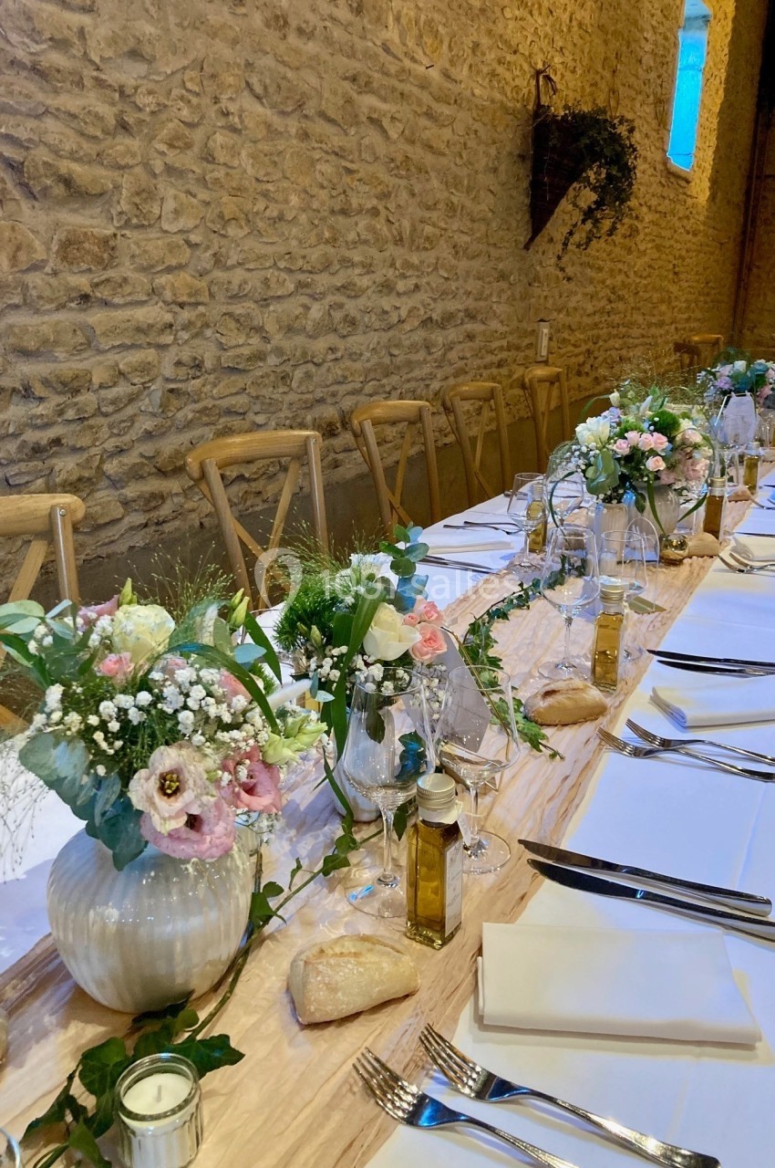 Table de réception décorée avec des fleurs, nappes blanches, vaisselle élégante et murs en pierre apparente.
