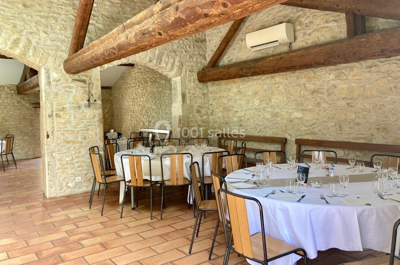 Salle en pierre avec poutres apparentes, tables rondes dressées avec nappes blanches et chaises en bois.