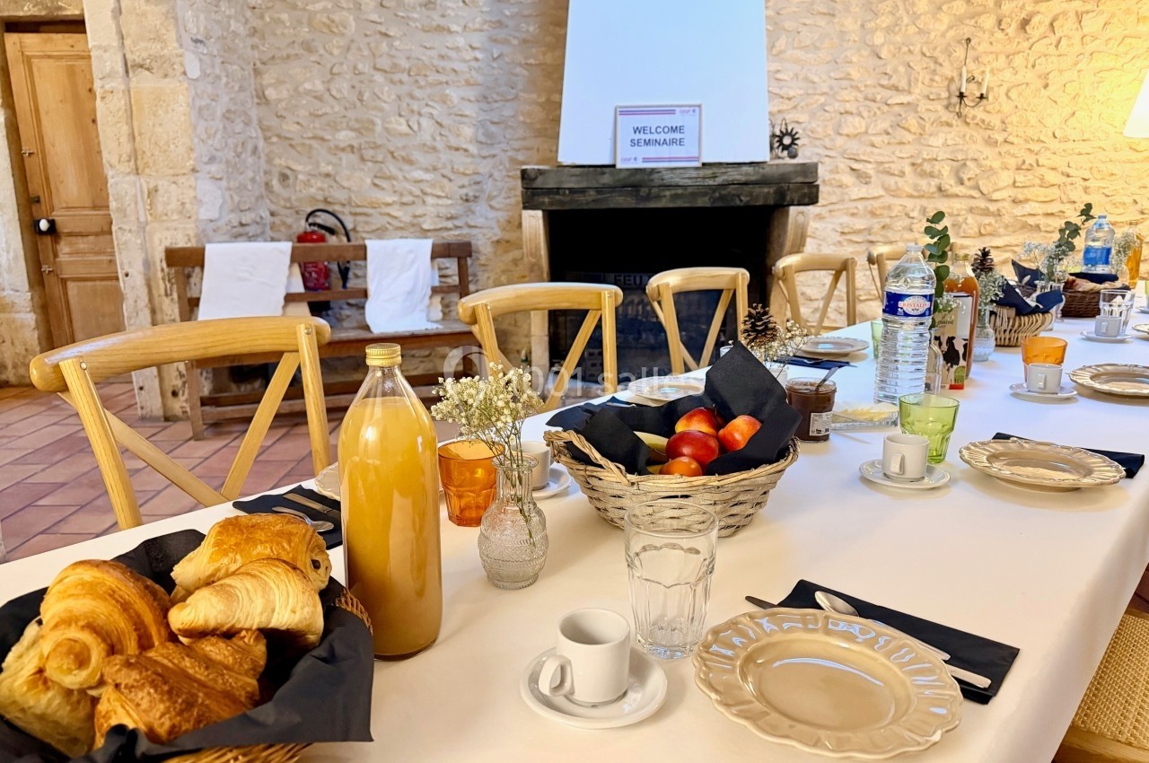 Table dressée avec viennoiseries, jus, fruits et boissons dans une salle en pierre avec cheminée en arrière-plan.