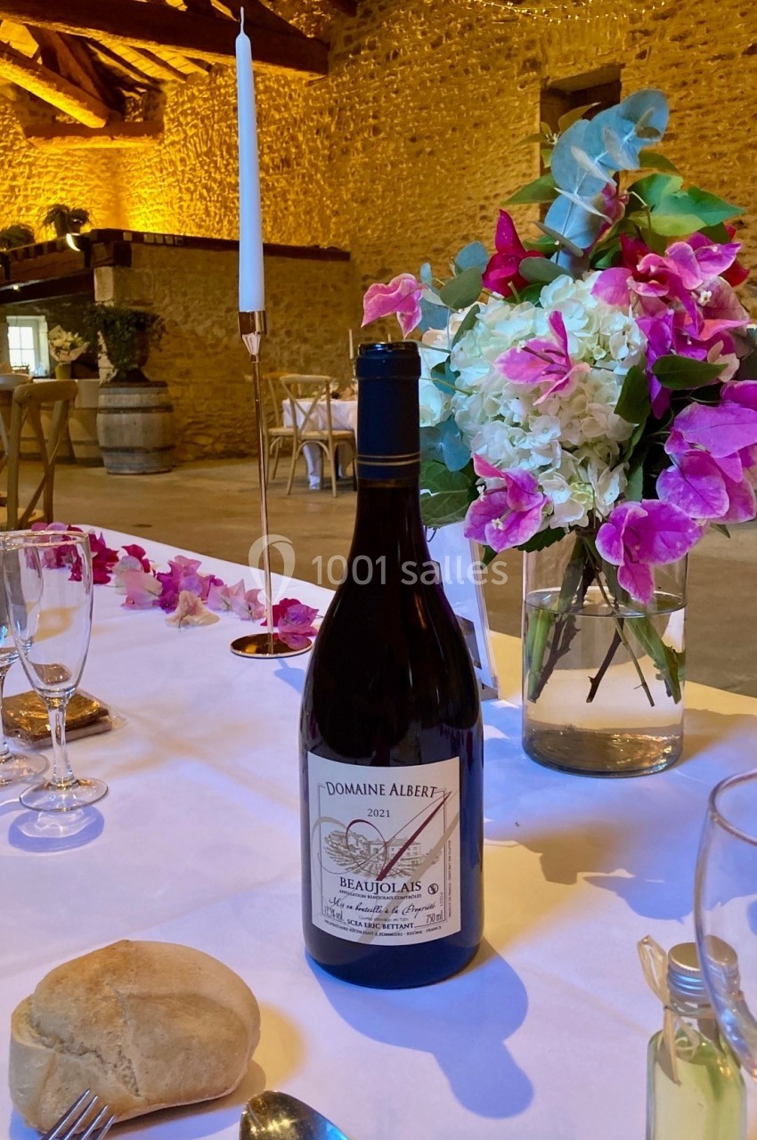 Bouteille de vin rouge sur une table dressée avec fleurs, bougie, verres et pain, dans une salle en pierre éclairée.