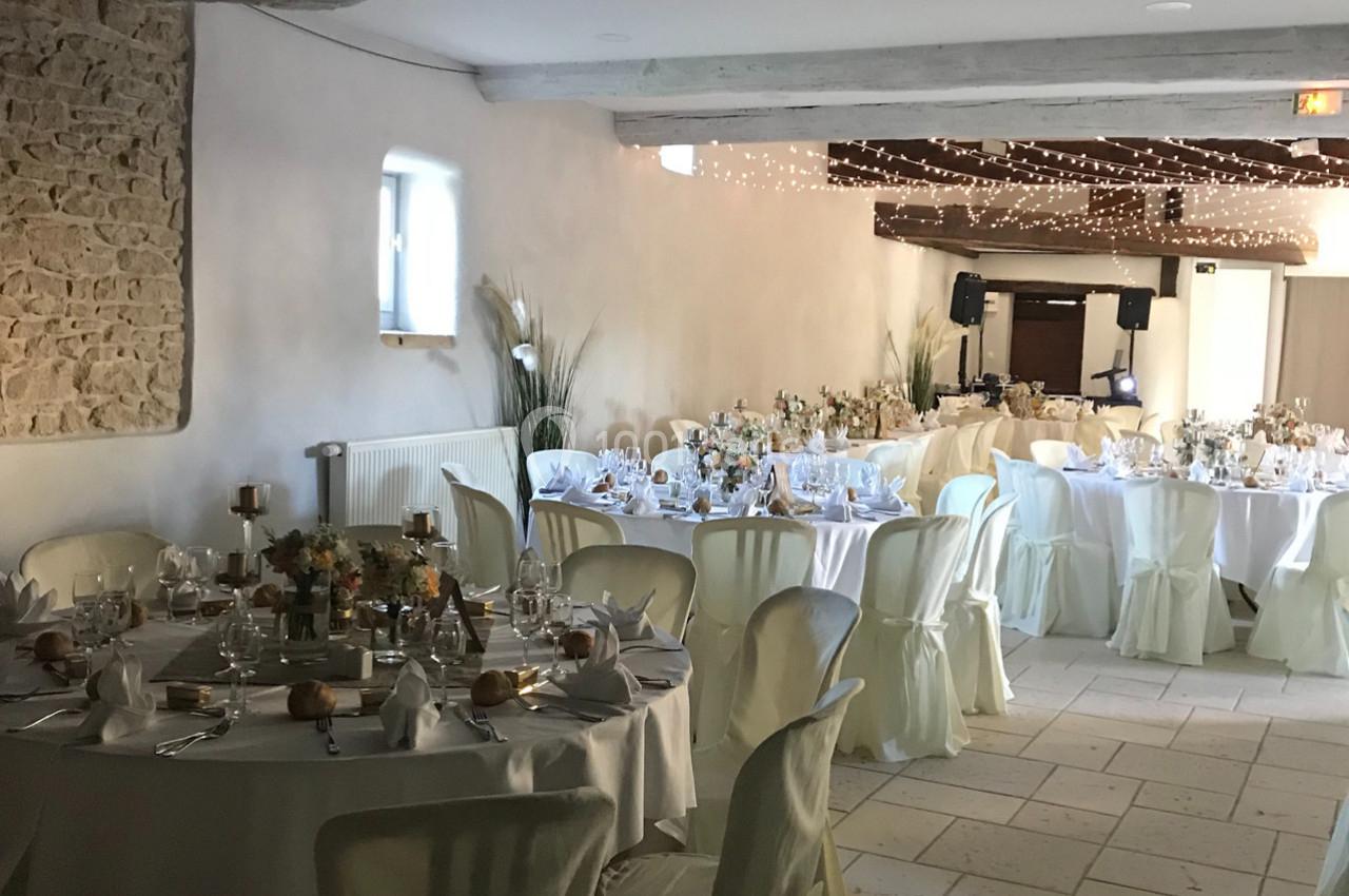 Salle de réception décorée pour un événement, avec tables rondes dressées, nappes blanches et guirlandes lumineuses au…