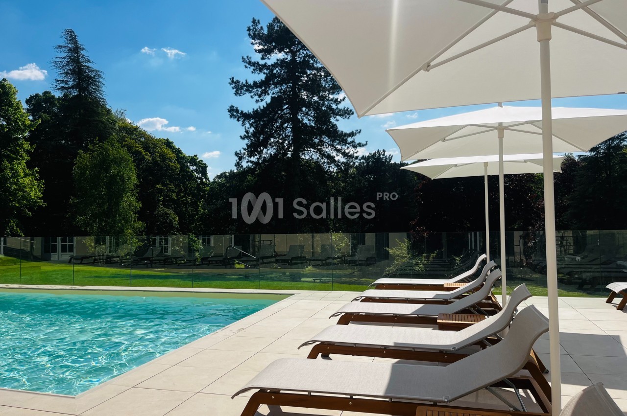 Chaises longues alignées sous des parasols blancs au bord d'une piscine, entourée de verdure et d'arbres.
