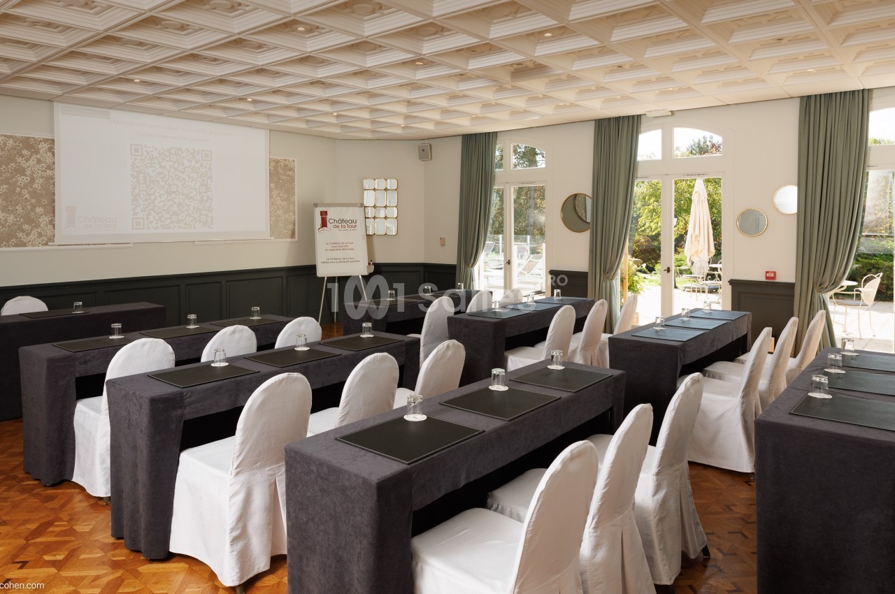 Salle de réunion lumineuse avec des tables alignées, chaises blanches et écran de projection à l'avant.