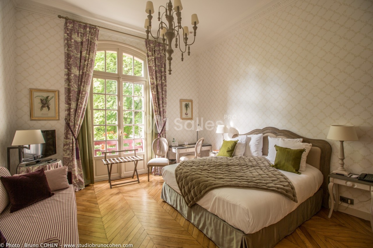 Chambre lumineuse avec lit double, fauteuils, bureau et grande fenêtre donnant sur un jardin verdoyant.