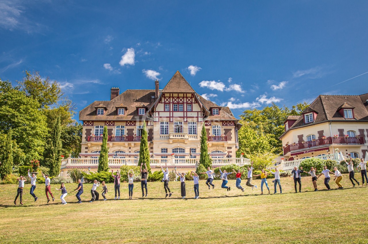 Un groupe de personnes saute en ligne devant un grand bâtiment à colombages entouré de verdure par temps ensoleillé.