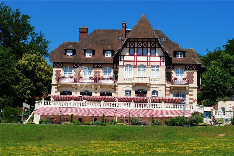 Façade d'un grand bâtiment à colombages avec balcons, entouré de verdure et sous un ciel bleu.