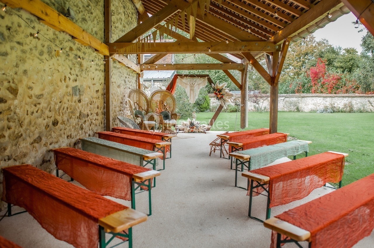 Espace de cérémonie en plein air sous une pergola en bois, avec bancs décorés de tissus colorés et vue sur un jardin.