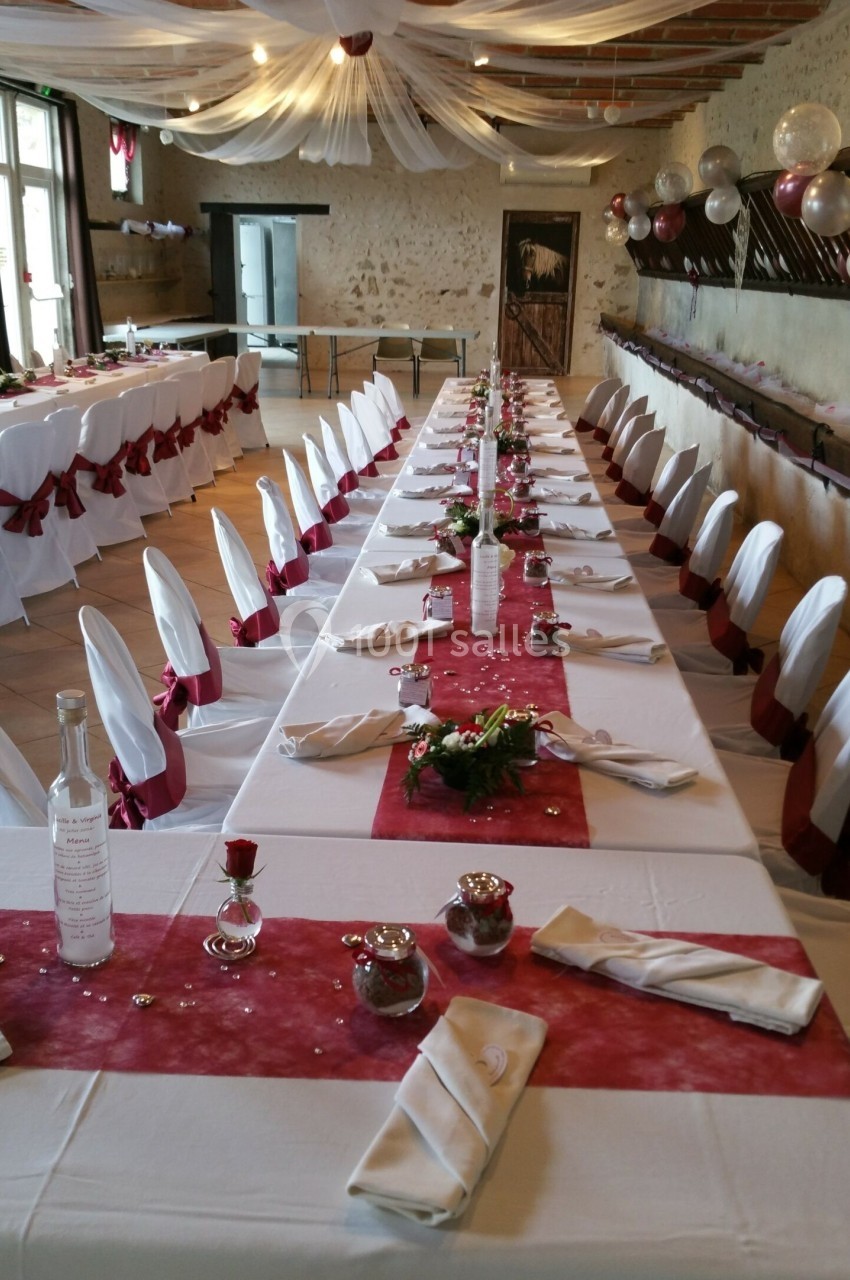 Salle de réception décorée avec des tables alignées, nappes blanches, chemins de table rouges et chaises ornées de nœuds.