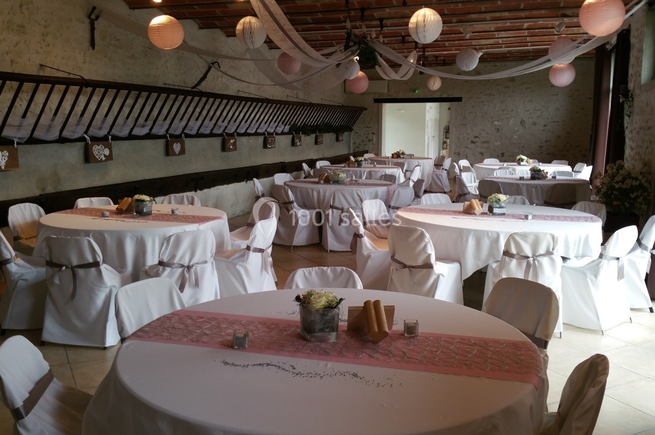 Salle de réception décorée avec des tables rondes, nappes blanches, chemins de table roses et lanternes suspendues.