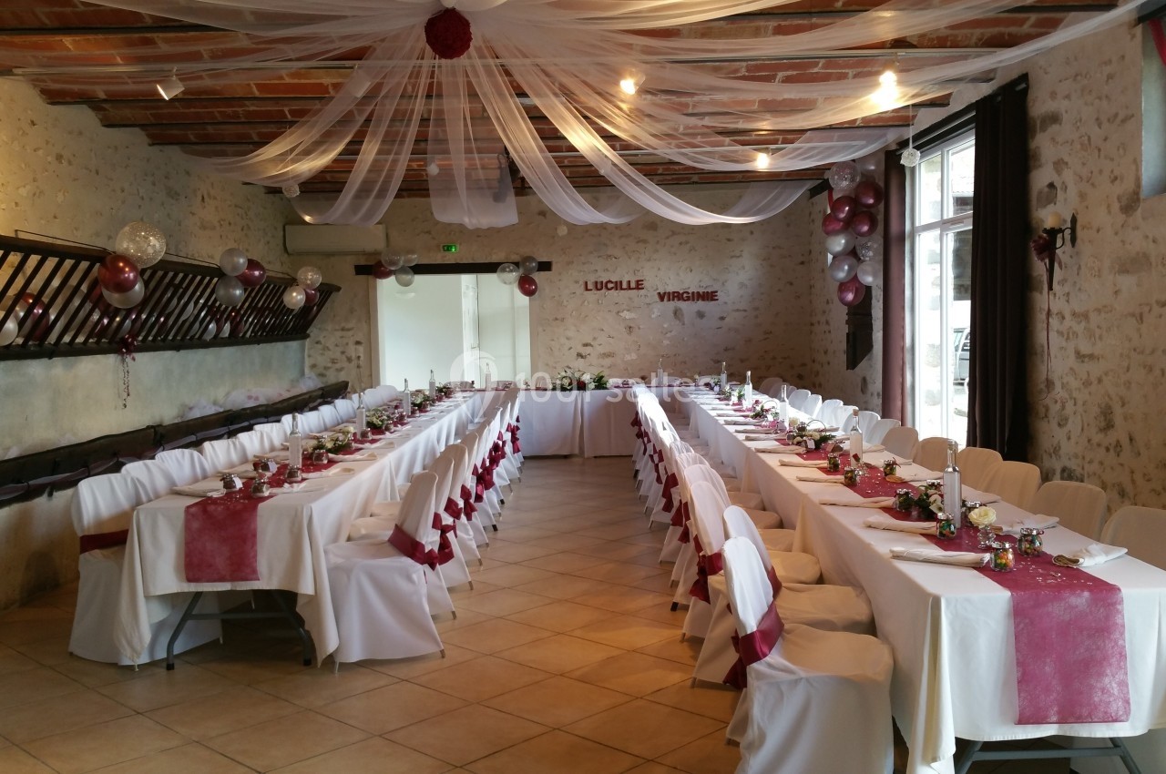 Salle de réception décorée avec des tables alignées, nappes blanches, chemins de table rouges et guirlandes suspendues.