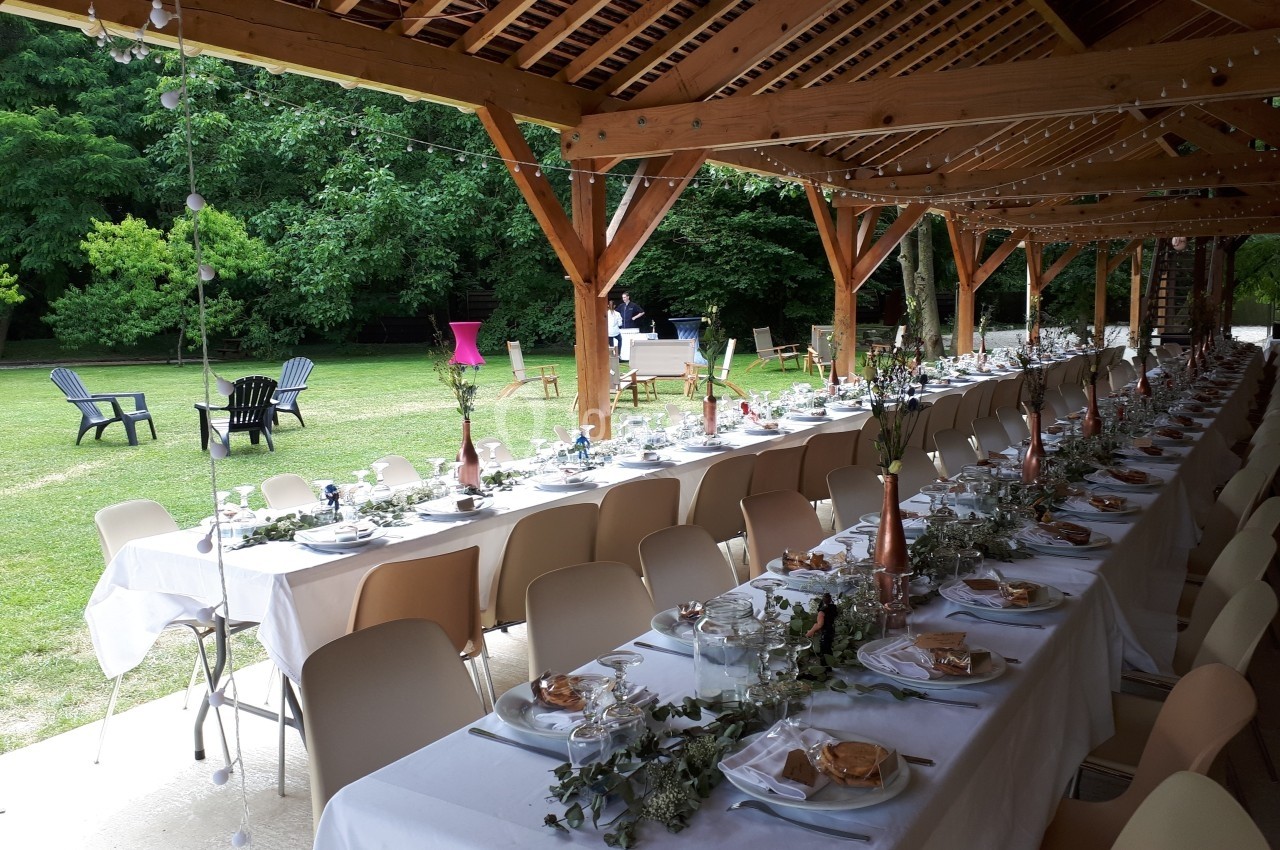 Tables dressées pour un repas en extérieur sous une pergola en bois, avec vue sur un jardin verdoyant.