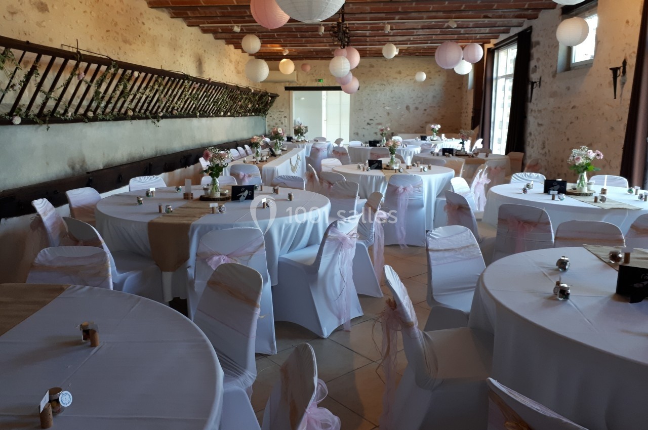 Salle de réception décorée avec des tables rondes, nappes blanches, chaises ornées de rubans roses et lanternes suspendues.