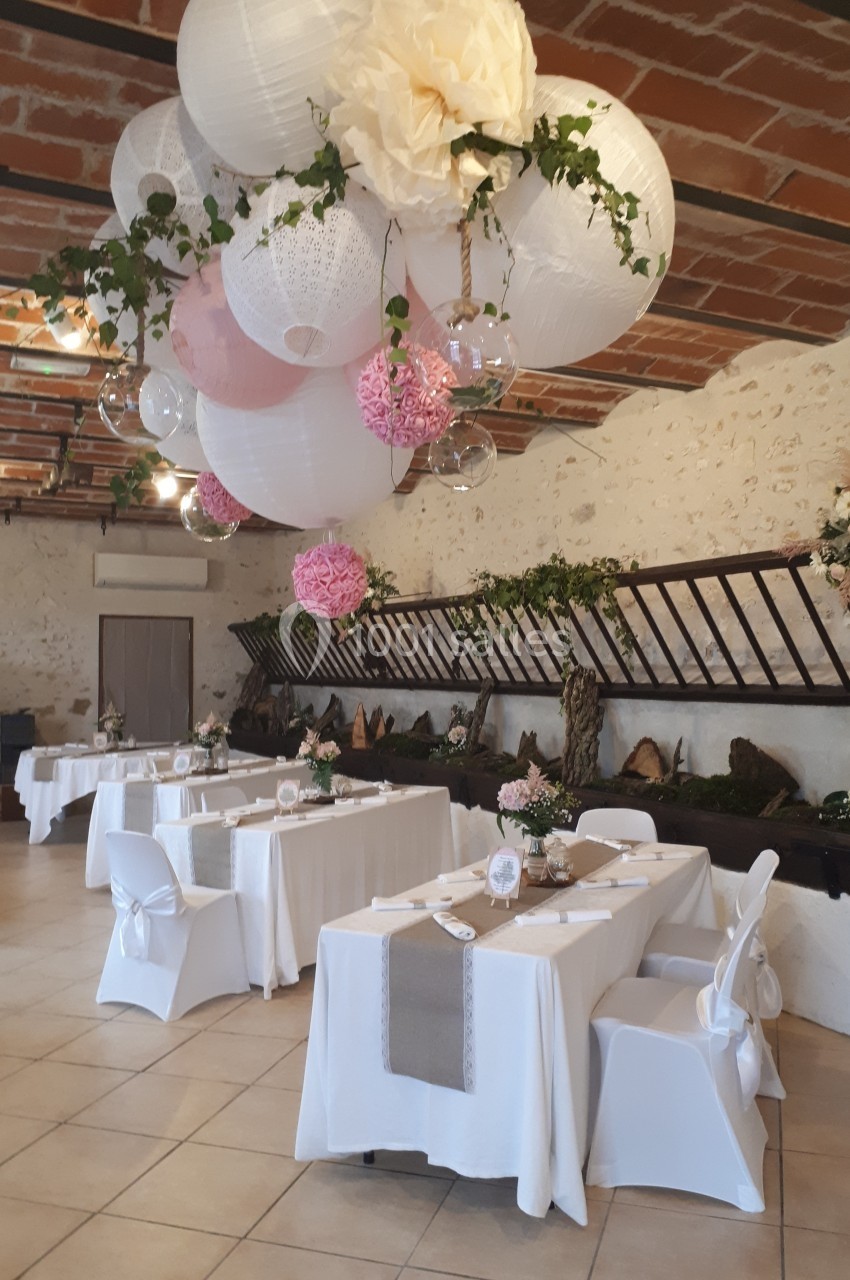 Salle de réception décorée avec des lanternes suspendues, des fleurs roses et blanches, et des tables élégamment dressées.