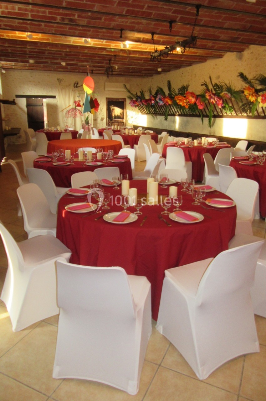 Salle décorée pour un événement avec des tables rondes nappées de rouge, chaises blanches et centre de table avec bougies.