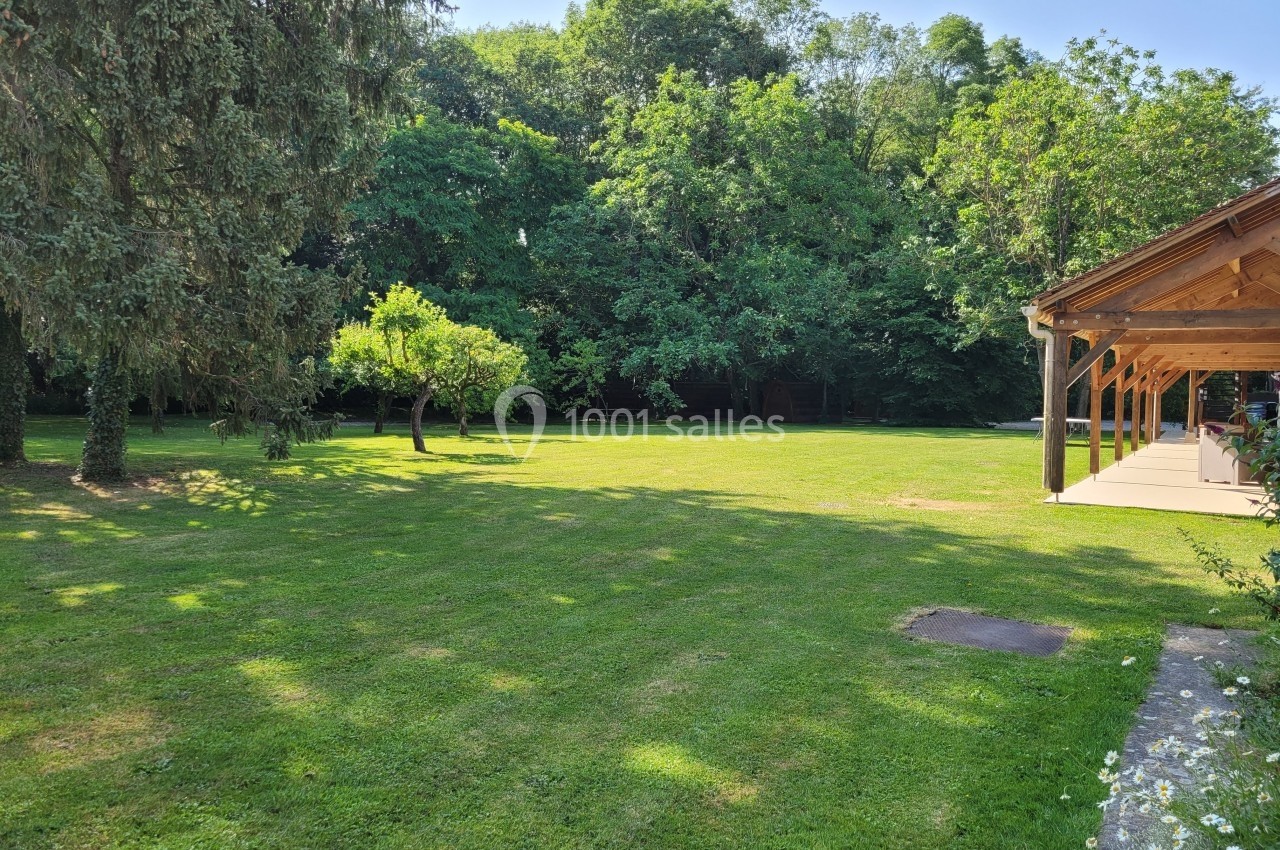 Jardin verdoyant avec pelouse, arbres et terrasse en bois sur la droite, bordé par une forêt en arrière-plan.