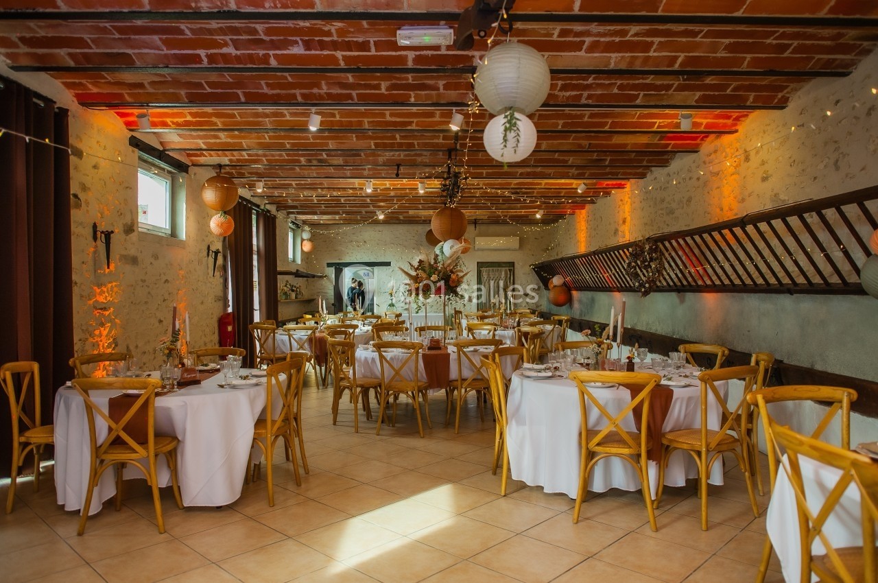 Salle de réception décorée avec des tables rondes dressées, des chaises en bois et des lanternes suspendues.