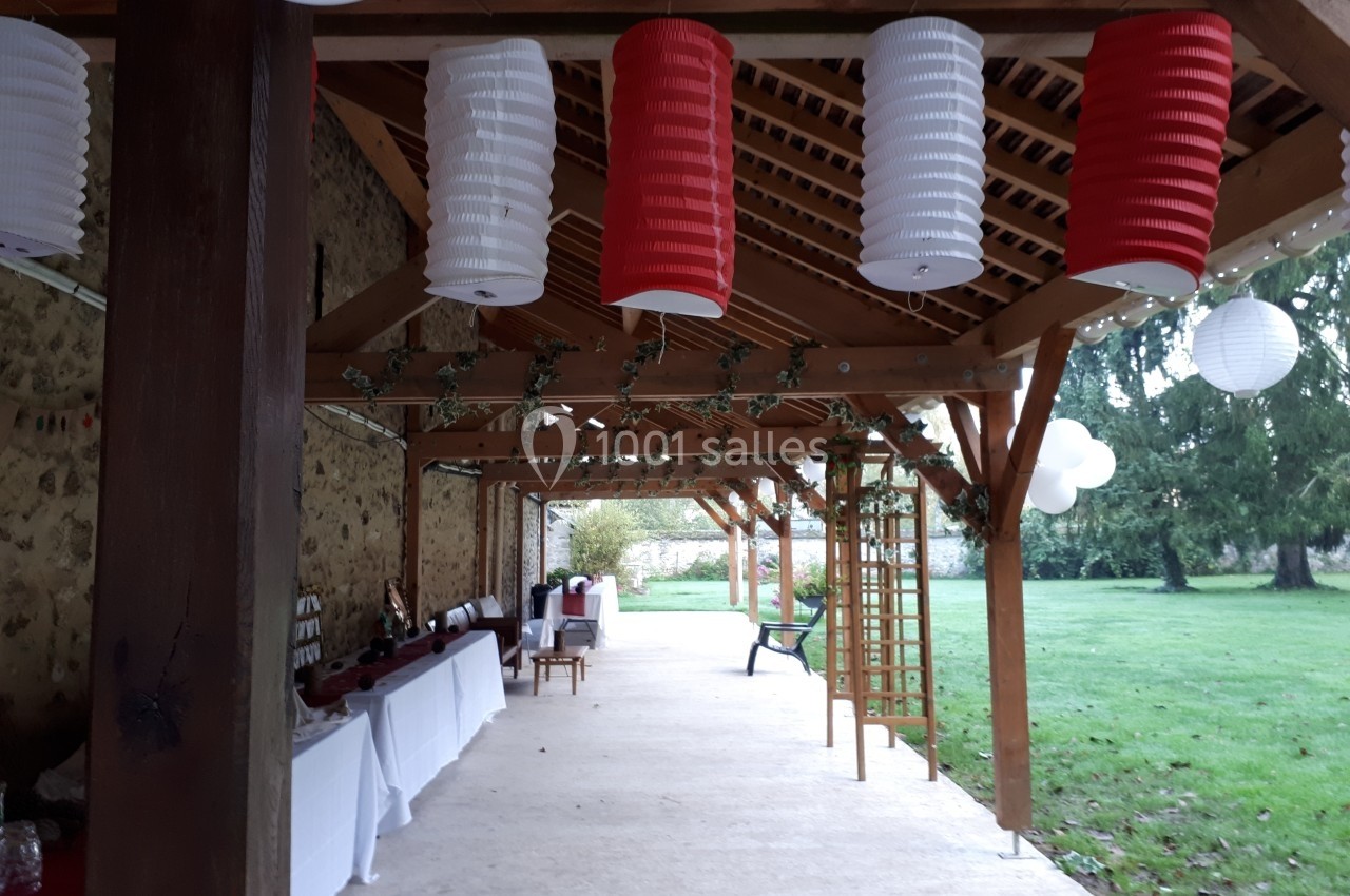 Pergola en bois décorée de lanternes rouges et blanches, tables dressées sur le côté, donnant sur un jardin verdoyant.