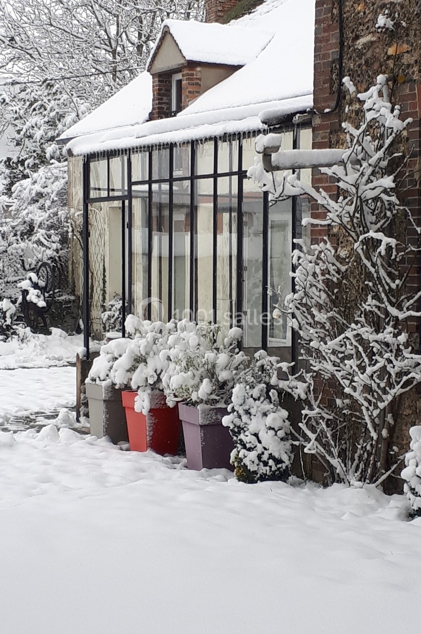 Façade d'une maison en hiver avec véranda, pots de fleurs enneigés et jardin couvert de neige.