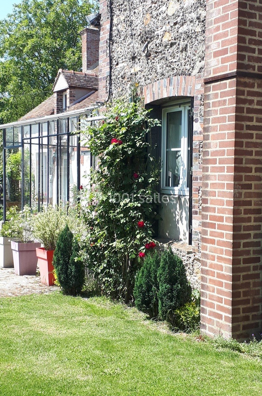 Façade en pierre et brique d'une maison avec rosiers grimpants, jardin verdoyant et terrasse ensoleillée.