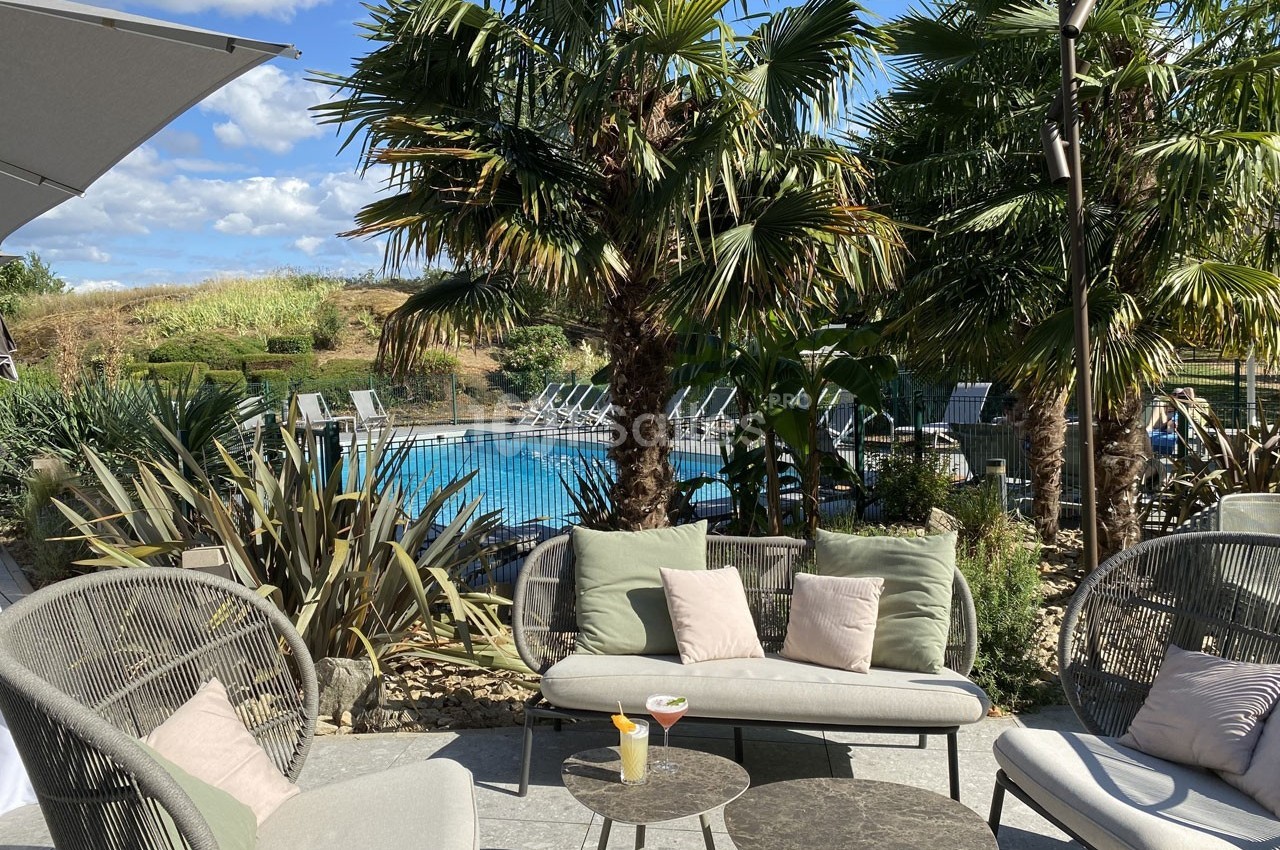 Terrasse aménagée avec fauteuils et table basse, vue sur une piscine entourée de palmiers et végétation.