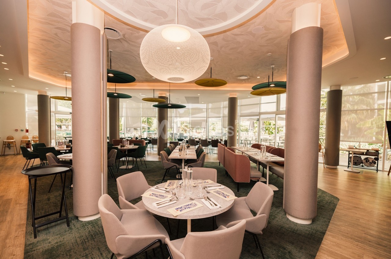 Salle de restaurant lumineuse avec tables dressées, chaises modernes et grandes baies vitrées donnant sur l'extérieur.
