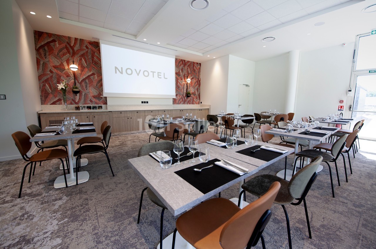 Salle de restaurant lumineuse avec tables dressées, chaises modernes et écran de projection au fond.