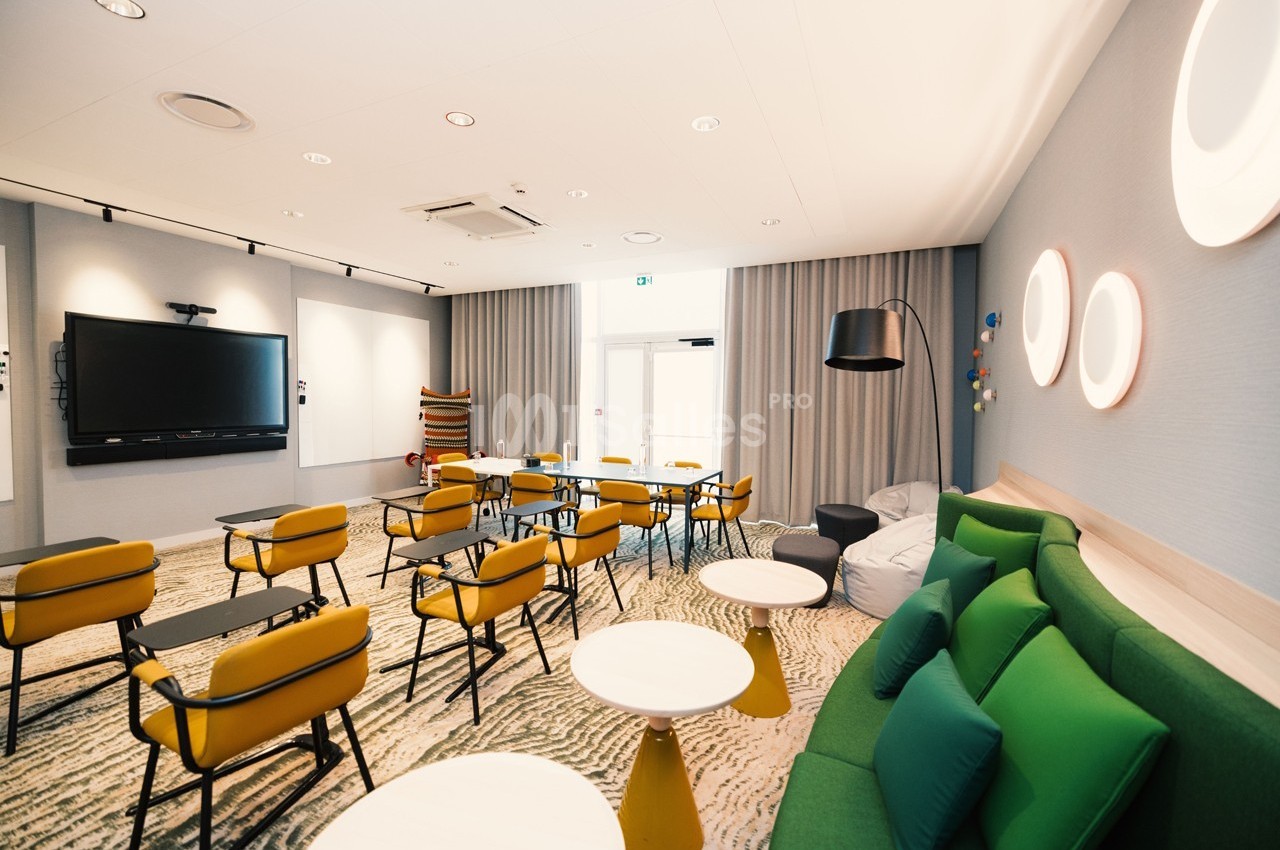 Salle de réunion lumineuse avec chaises jaunes, tables noires, canapé vert et écran mural.