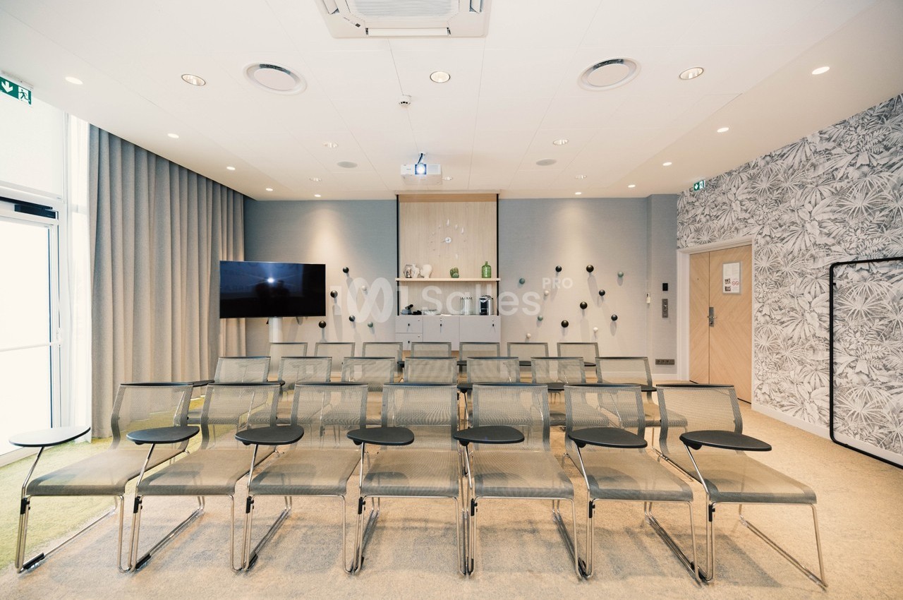 Salle de réunion lumineuse avec rangées de chaises équipées de tablettes, écran et projecteur au fond.