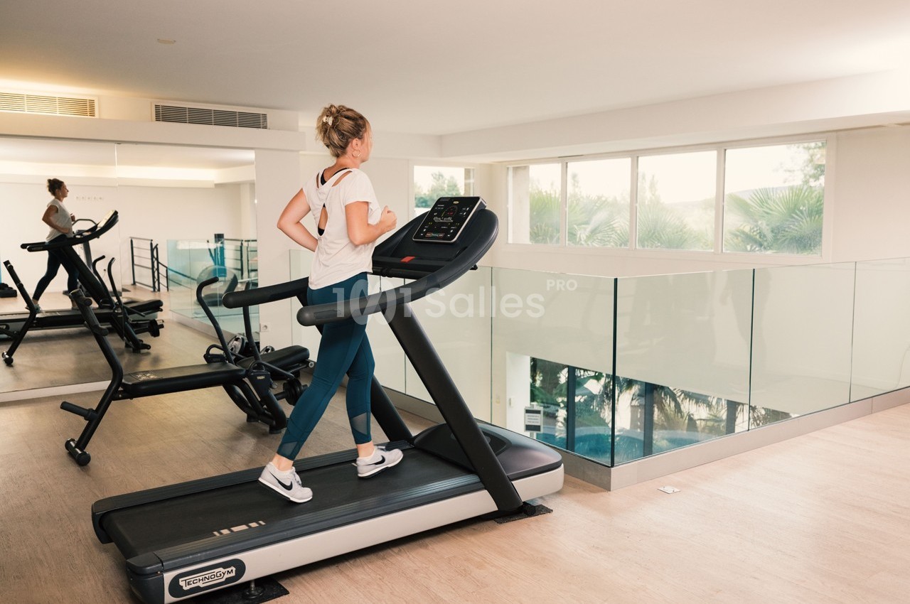 Femme courant sur un tapis de course dans une salle de sport lumineuse avec vue sur des arbres et une piscine.