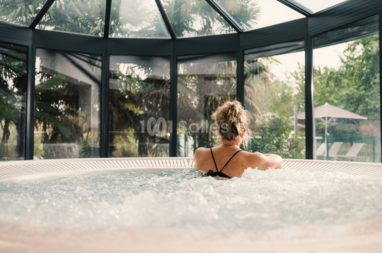 Femme de dos se relaxant dans un jacuzzi sous une verrière, avec vue sur un jardin verdoyant.