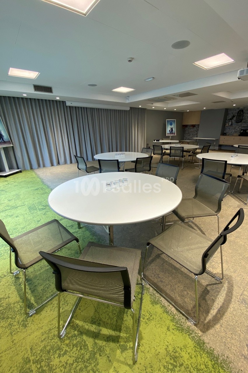 Salle de réunion lumineuse avec tables rondes blanches, chaises noires et sol mêlant moquette verte et béton.