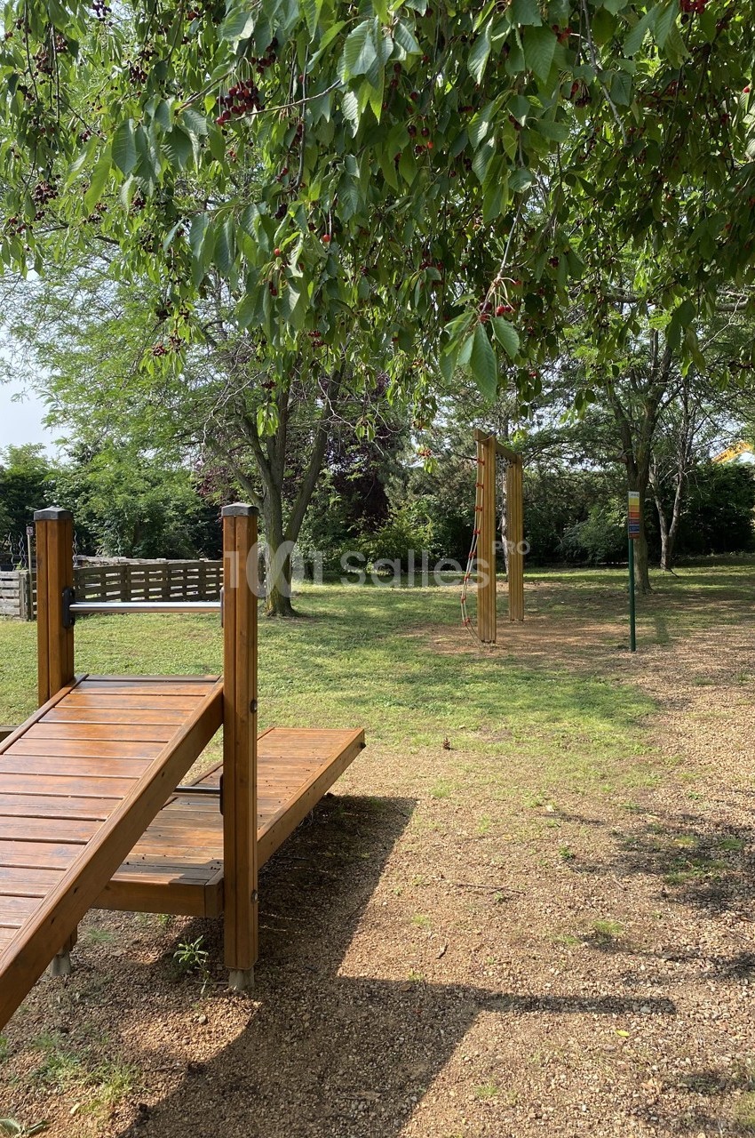 Structure en bois dans un parc verdoyant avec des arbres, un sol en gravier et des équipements de jeu en arrière-plan.