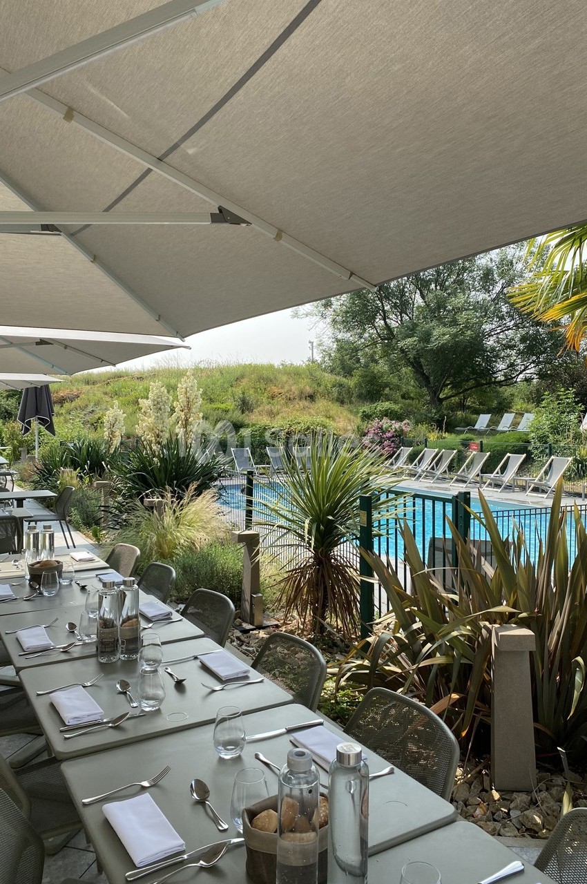 Terrasse ombragée avec tables dressées, vue sur une piscine entourée de végétation et de transats.