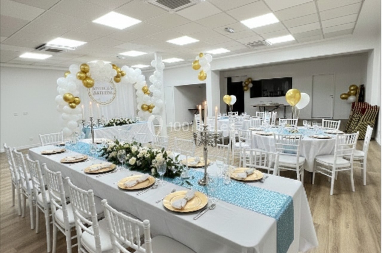 Salle décorée pour une réception avec tables dressées, nappes blanches, chandeliers et ballons dorés et blancs.