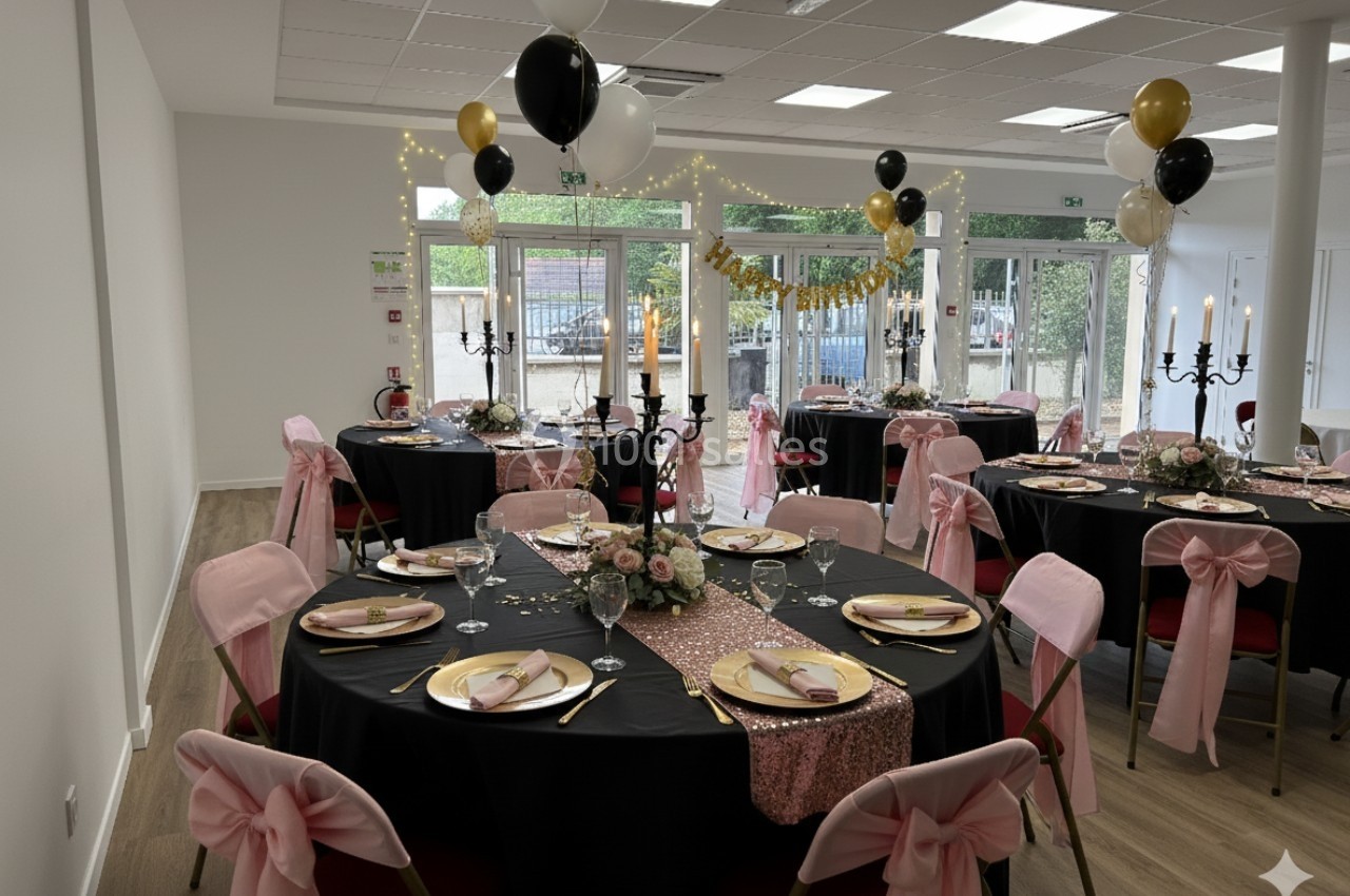 Salle décorée pour un événement avec tables rondes dressées, nappes noires, chaises ornées de nœuds roses et ballons…