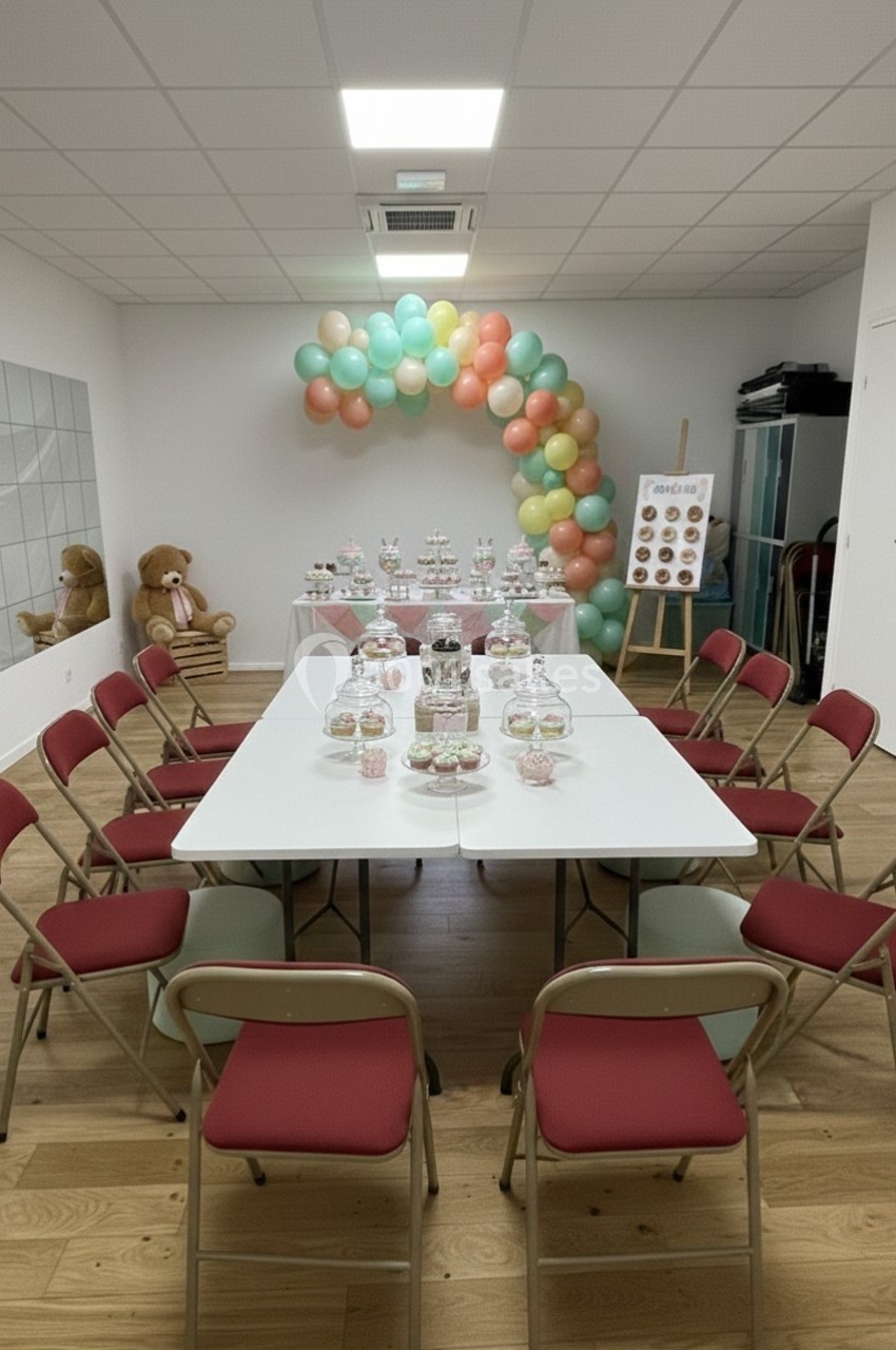 Salle décorée pour une fête, avec une table centrale entourée de chaises rouges et des ballons colorés en arrière-plan.